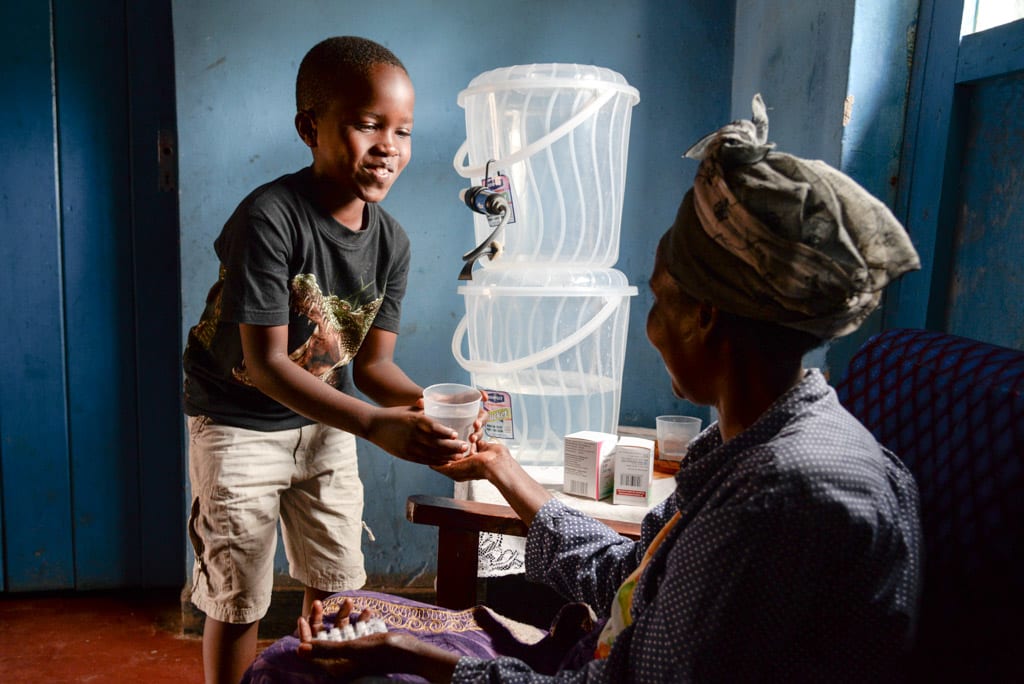8 year old Manzi Kevin, boy, child, wearing a black shirt and tan shorts, smiles as he is handing his mother, parent, adult woman, a cup of clean safe sanitary fresh purified drinking water in her hands, while in her other hands are her HIV medication and pills for her disease and illness. There is the clear stacked buckets, water filter and purification water containers, pails on the table beside them inside their home.